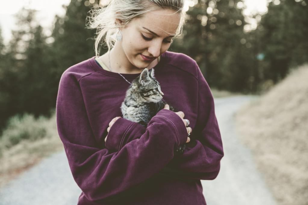 A blonde woman wearing a purple sweatshirt holds a tabby kitten in her arms.